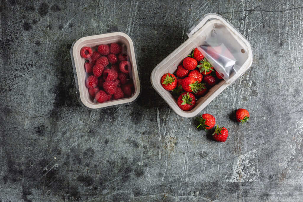 Punnet of strawberries and a punnet of raspberries in compostable packaging, on a grey background.