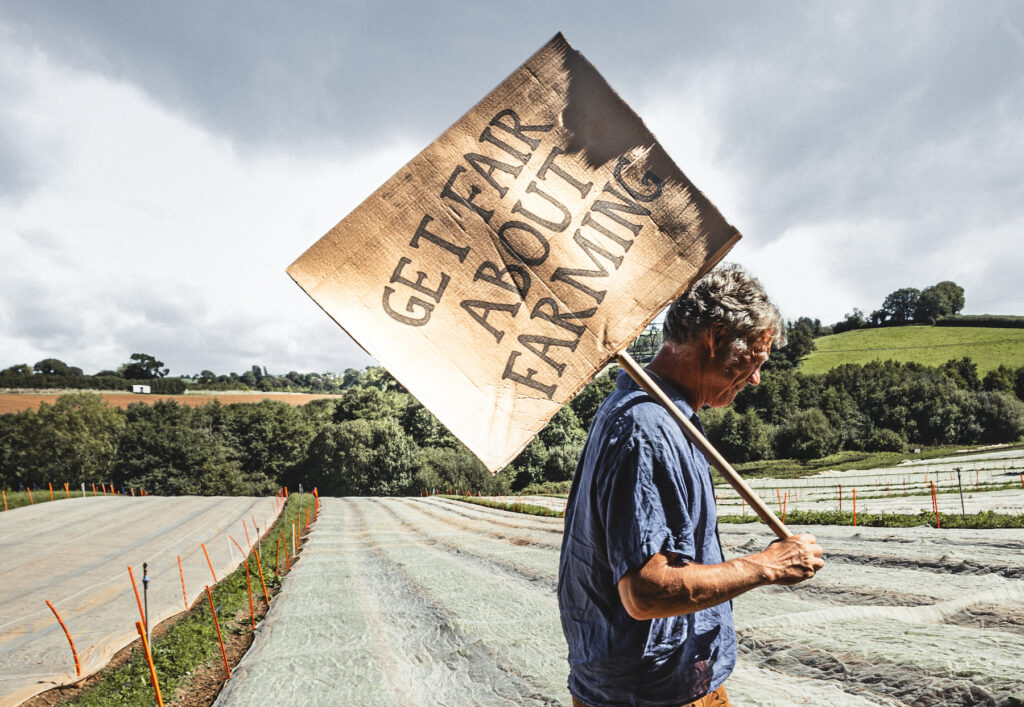 male farmer walking across a field carrying a sign that reads 'Get Fair About Farming'