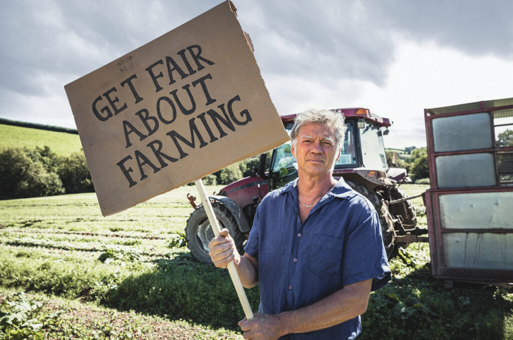 Guy Singh-Watson, founder of Riverford, holding a sign saying 'Get Fair About Farming' with a tractor and agricultural field in the background.